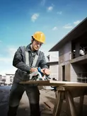 A person wearing safety equipment cuts wood with a cordless circular saw at a construction site.