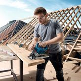 A person wearing safety equipment cuts wood with a cordless circular saw at a construction site.