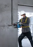 A person wearing safety equipment drills into concrete with a cordless rotary hammer.