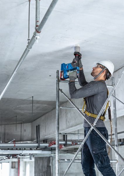 GBH 187-LI A person wearing safety equipment drills into a concrete ceiling while standing on scaffolding.