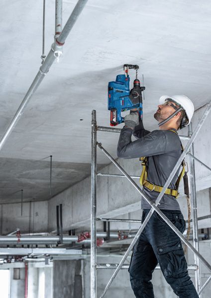GBH 187-LI A person wearing safety equipment drills into a concrete ceiling while standing on scaffolding.