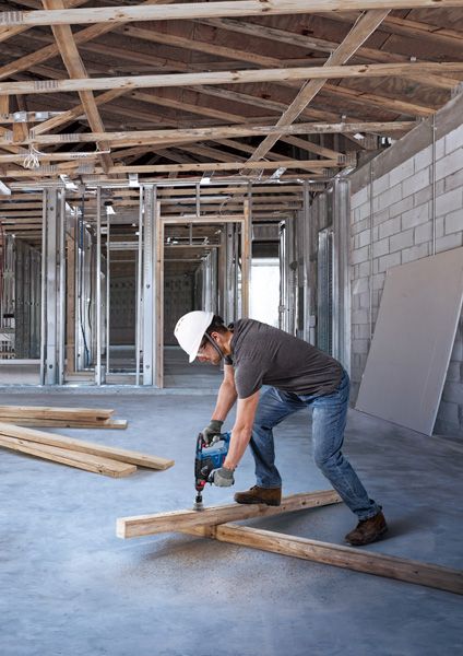 A person wearing safety equipment drills into a wooden beam at a construction site.