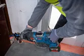 A person wearing safety equipment uses a reciprocating saw to cut metal near a door frame.