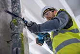 A person wearing safety equipment cuts a metal pipe with a reciprocating saw.