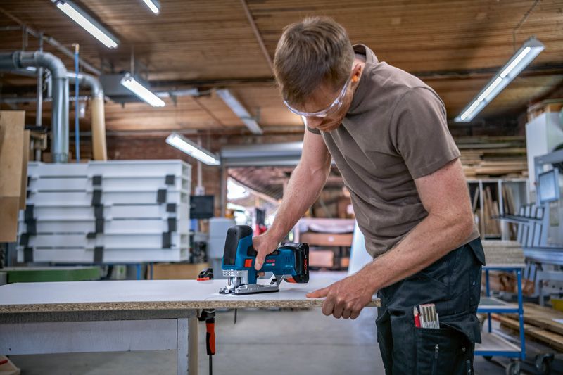 A person wearing safety equipment cuts a wood board with a jigsaw in a workshop.