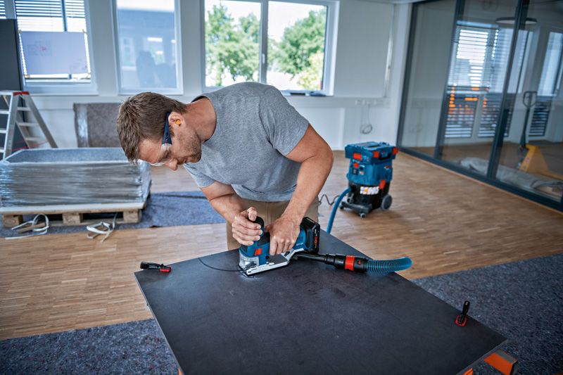 A person wearing safety equipment uses a jigsaw to cut a sheet on a workbench.