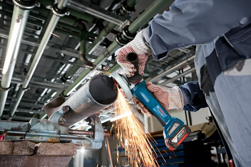 A person wearing safety equipment grinds a metal pipe with a cordless angle grinder.