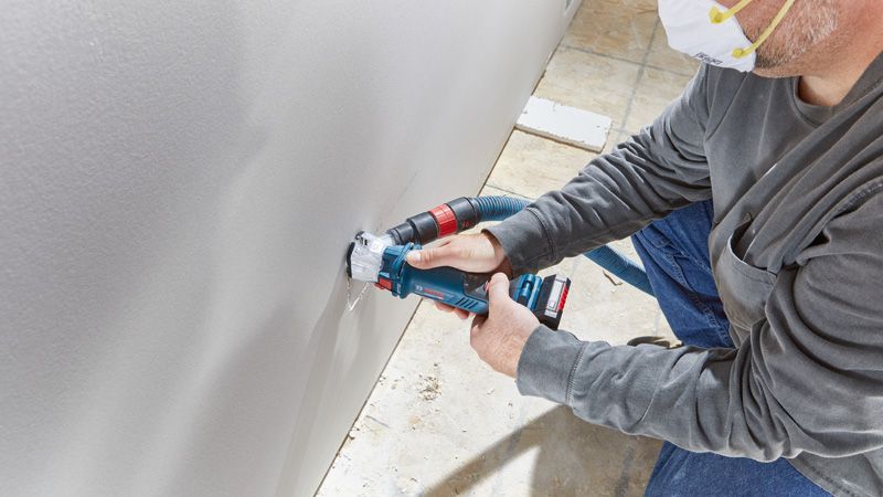 A person wearing safety equipment cuts a hole in drywall using a cordless drywall router.