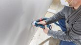 A person wearing safety equipment cuts a hole in drywall using a cordless drywall router.