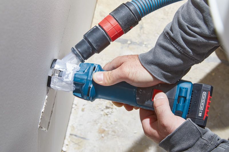 A person wearing safety equipment uses a cordless drywall router with dust extraction.