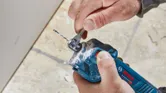 Person adjusts a cordless drywall router bit using a wrench near a drywall surface.
