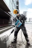 A person wearing safety equipment operates a breaker to demolish concrete at a construction site.