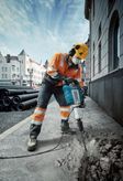 A person wearing safety equipment breaks pavement with a power breaker on a city street.