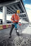 A person wearing safety equipment breaks concrete with a large electric breaker outdoors.