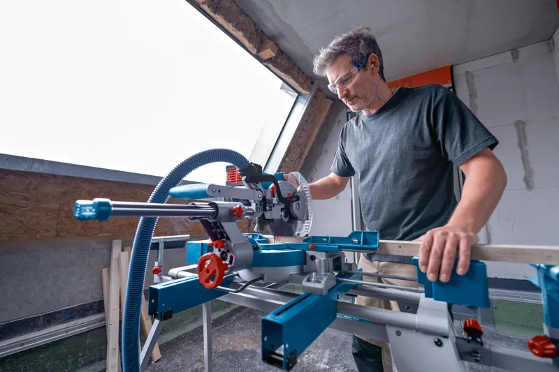 Person wearing safety equipment uses a cordless mitre saw to cut wood indoors.