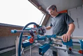 Person wearing safety equipment uses a cordless mitre saw to cut wood indoors.