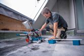 A person wearing safety equipment cuts a board with a cordless mitre saw on a rooftop.