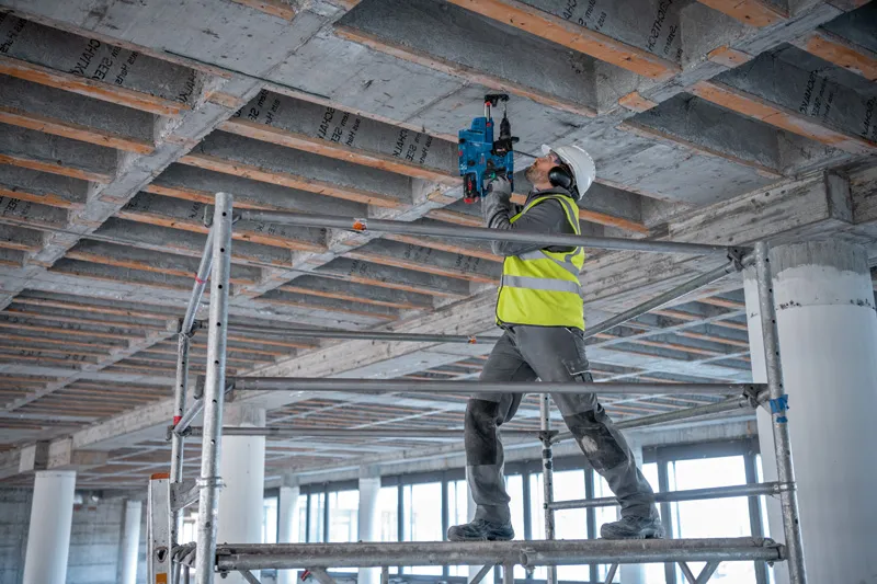GBH 18V-28 CF A person wearing safety equipment drills into a concrete ceiling at a construction site.