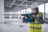 A person wearing safety equipment drills into concrete using a cordless rotary hammer.