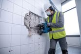 Person wearing safety equipment uses a cordless rotary hammer to break through a tiled wall.