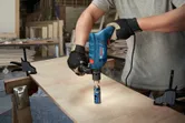 A person wearing safety equipment drills a hole into wood using an impact drill.