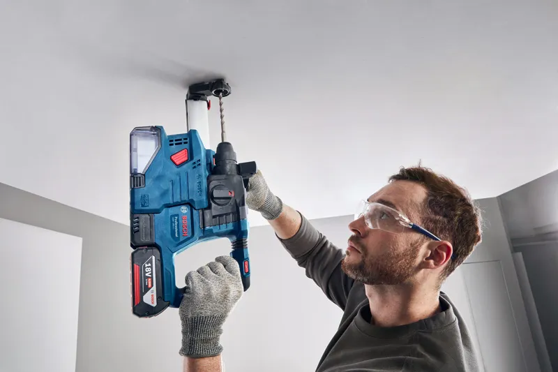 GDE 12 A person wearing safety equipment drills into a ceiling using a cordless rotary hammer.