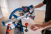 A person uses a cordless mitre saw to cut wood, guided by a laser leveling tool.