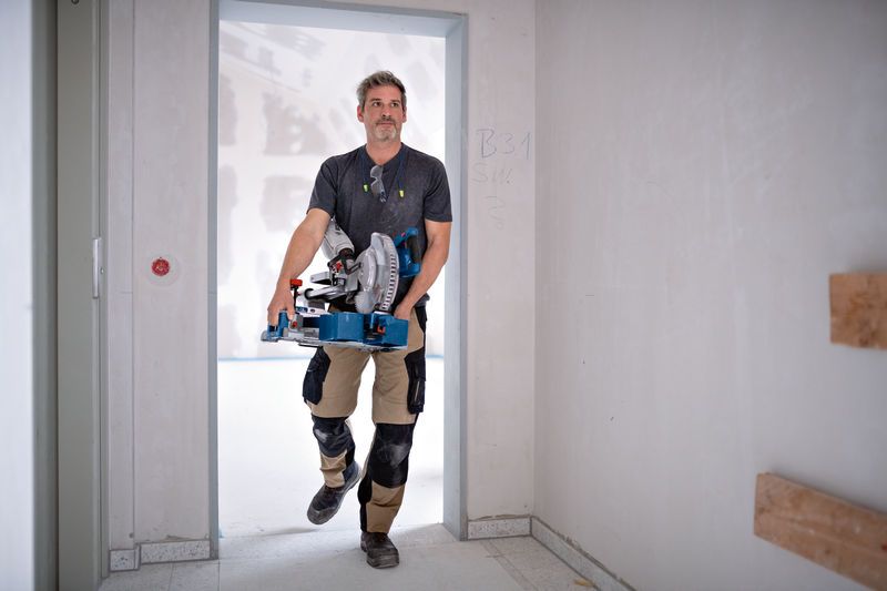 A person wearing safety equipment carries a cordless mitre saw through a doorway.