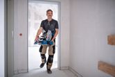 A person wearing safety equipment carries a cordless mitre saw through a doorway.
