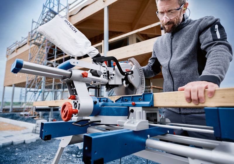 A person wearing safety equipment cuts wood with a cordless mitre saw at a construction site.