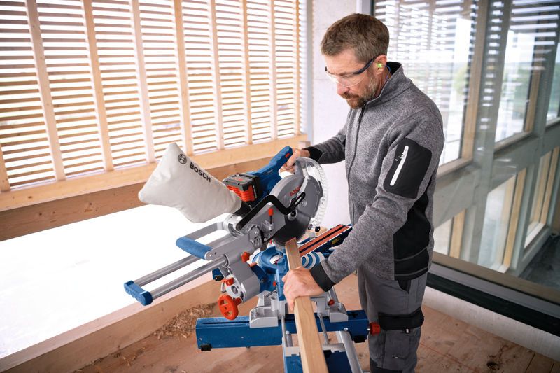 A person wearing safety equipment operates a cordless mitre saw to cut wooden trim.