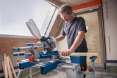 A person wearing safety equipment operates a cordless mitre saw to cut wood indoors.