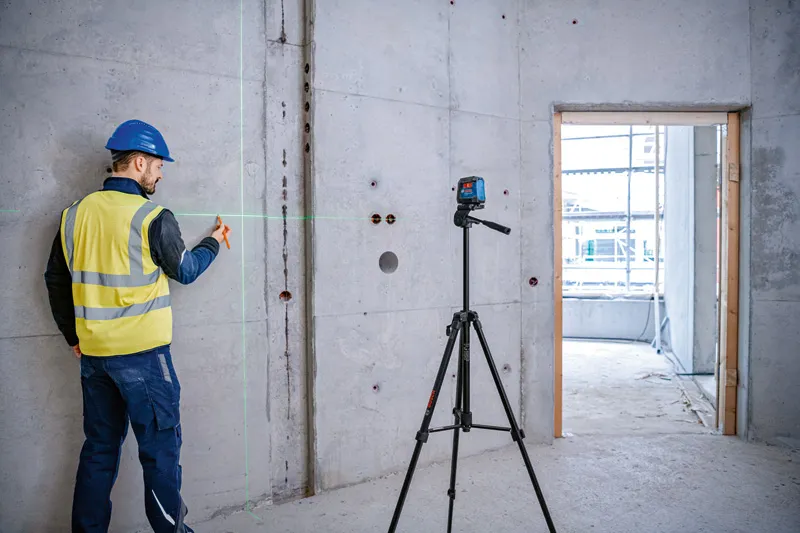 A person wearing safety equipment marks a concrete wall using a laser leveling tool on a tripod.