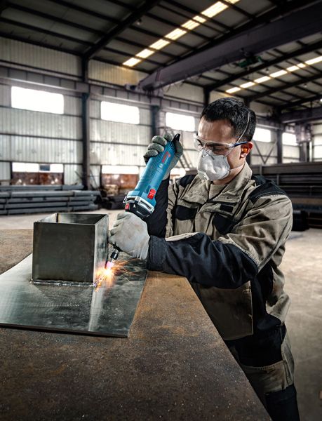 A person wearing safety equipment uses a straight grinder on a metal sheet.