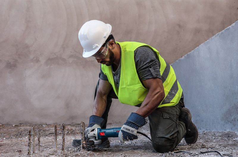 A person wearing safety equipment cuts exposed rebar with an angle grinder.