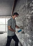 A person wearing safety equipment uses an angle grinder to cut into a concrete wall.