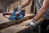 A person wearing safety equipment planes a wooden plank with a cordless tool.