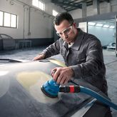 A person wearing safety equipment uses a random orbit sander on a car hood.