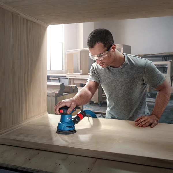 GEX 125 A person wearing safety equipment sands wood with a random orbit sander inside a workshop.