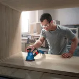 A person wearing safety equipment sands wood with a random orbit sander inside a workshop.