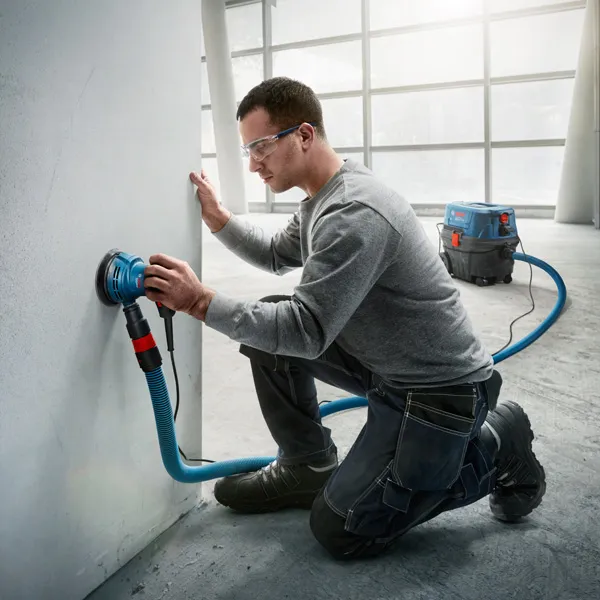 GEX 125 A person wearing safety equipment sands a wall using a random orbit sander with dust extraction.