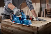 A person wearing safety equipment smooths wooden boards with a handheld planer.
