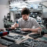 A person assembles electronics at a workbench using a cordless screwdriver.