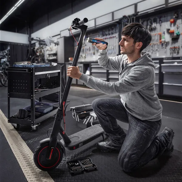 A person kneels and uses a cordless screwdriver to repair an electric scooter.