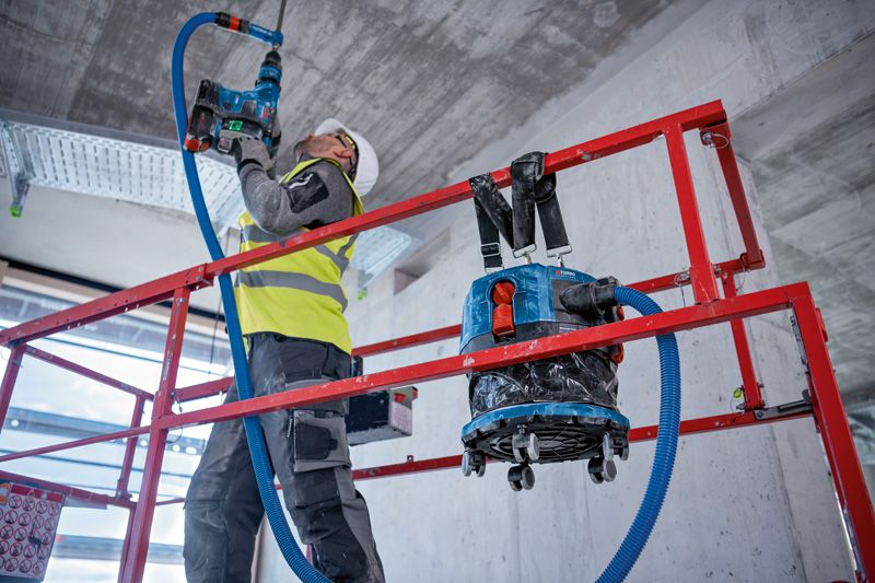 ProCORE18V+ 8.0Ah A person wearing safety equipment drills into a ceiling on a scaffold with a dust extractor nearby.