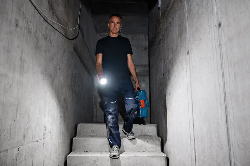 A person holds a cordless work light and tool case while descending concrete stairs.