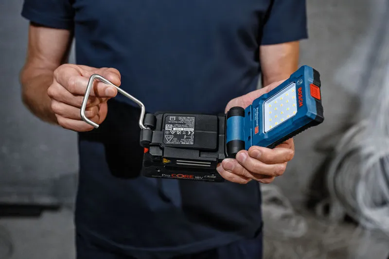 Person holding a cordless LED work light with a battery and metal handle.
