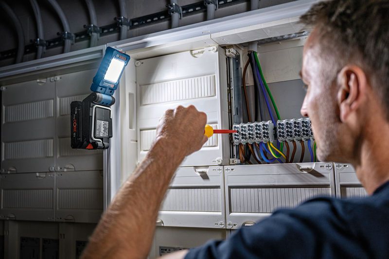 A person uses a screwdriver on an electrical panel, cordless light attached for illumination.