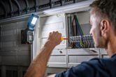 A person uses a screwdriver on an electrical panel, cordless light attached for illumination.