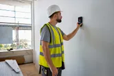 A person wearing safety equipment uses a wall detector on a white surface.